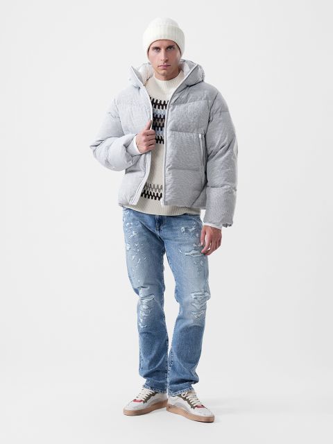 Un hombre que lleva una chaqueta gris claro, un jersey estampado, un gorro blanco y vaqueros azules desgastados, así como zapatillas deportivas.