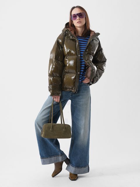 Una persona con una brillante chaqueta de plumas verde oliva, una camisa a rayas azules, jeans anchos y gafas de sol moradas, sosteniendo un bolso de mano verde oliva.