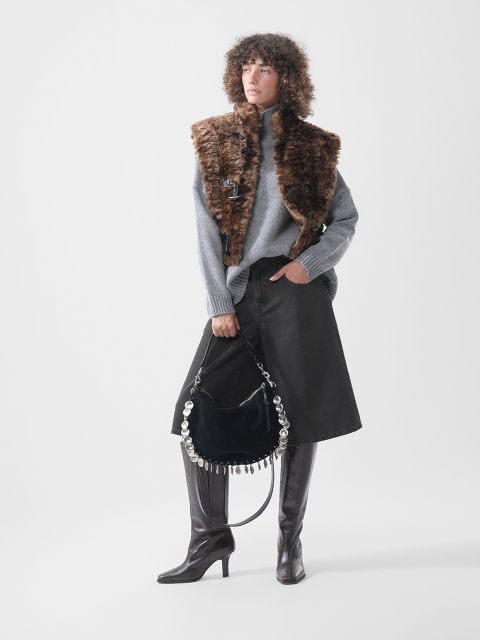 Una modelo lleva un jersey de cuello alto gris, un chaleco de piel sintética marrón y una falda vaquera negra; además, sostiene un bolso de mano negro con herrajes redondos plateados y lleva botas altas marrones.