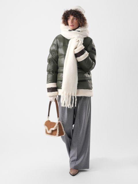Una persona con una chaqueta de plumas verde oliva con ribete de piel de cordero, una bufanda y un gorro blancos, pantalones grises y un pequeño bolso marrón con ribete de piel de cordero.