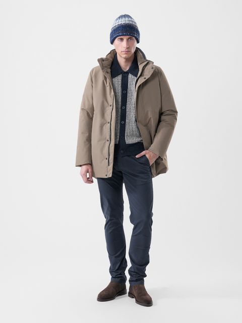 Un hombre que lleva una parka de color canela sobre un suéter de camisa estampada, pantalones oscuros, botas marrones y un gorro de punto a rayas.
